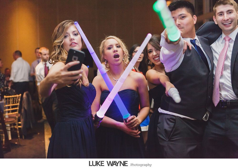 People at party pose with lightsabers, one taking selfie. Dark dresses, suits, glowing sticks, happy expressions.