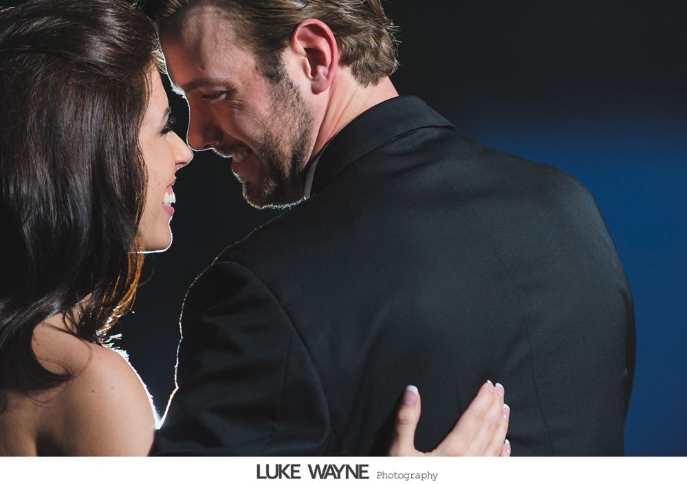 Couple embracing and smiling, man in black suit, woman in formal dress. Blue backdrop.
