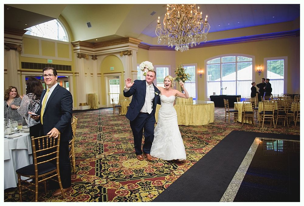 Bride with arms up, making a funny face, groom looking at his hand, cake cutting in background.