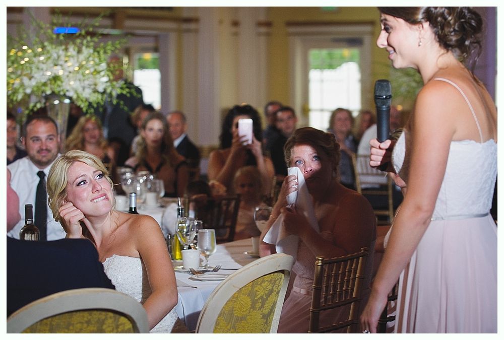 Bride with arms up, making a funny face, groom looking at his hand, cake cutting in background.