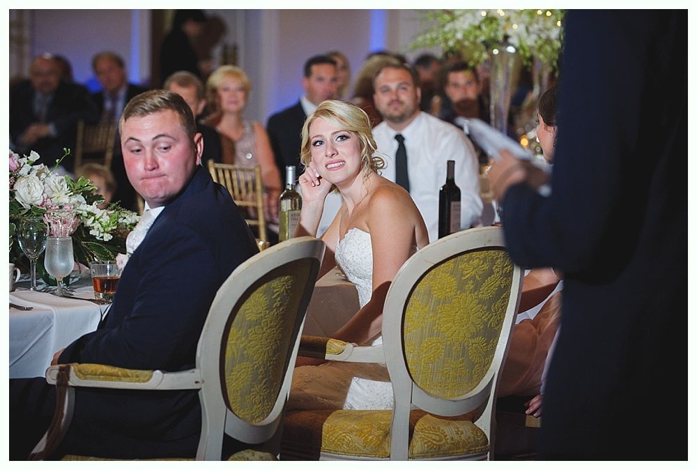 Bride with arms up, making a funny face, groom looking at his hand, cake cutting in background.