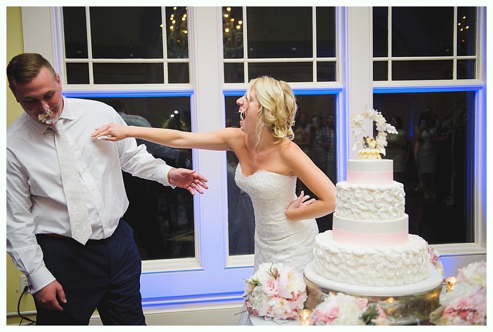 Bride with arms up, making a funny face, groom looking at his hand, cake cutting in background.