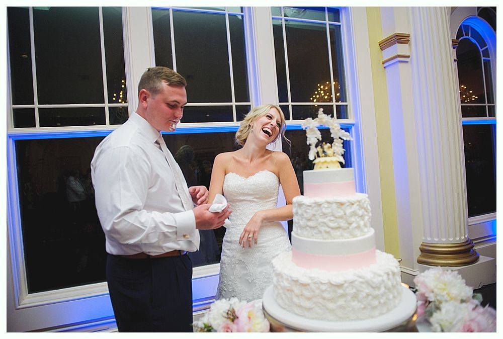 Bride with arms up, making a funny face, groom looking at his hand, cake cutting in background.