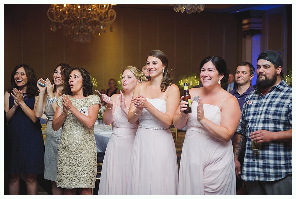 Bride with arms up, making a funny face, groom looking at his hand, cake cutting in background.