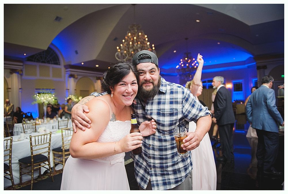 Bride with arms up, making a funny face, groom looking at his hand, cake cutting in background.