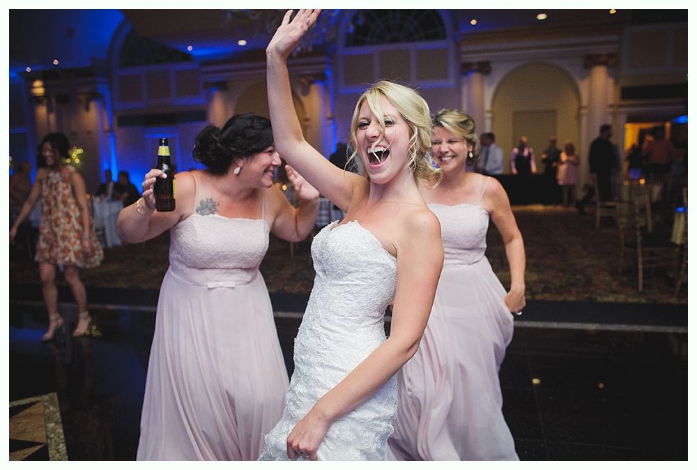 Bride with arms up, making a funny face, groom looking at his hand, cake cutting in background.