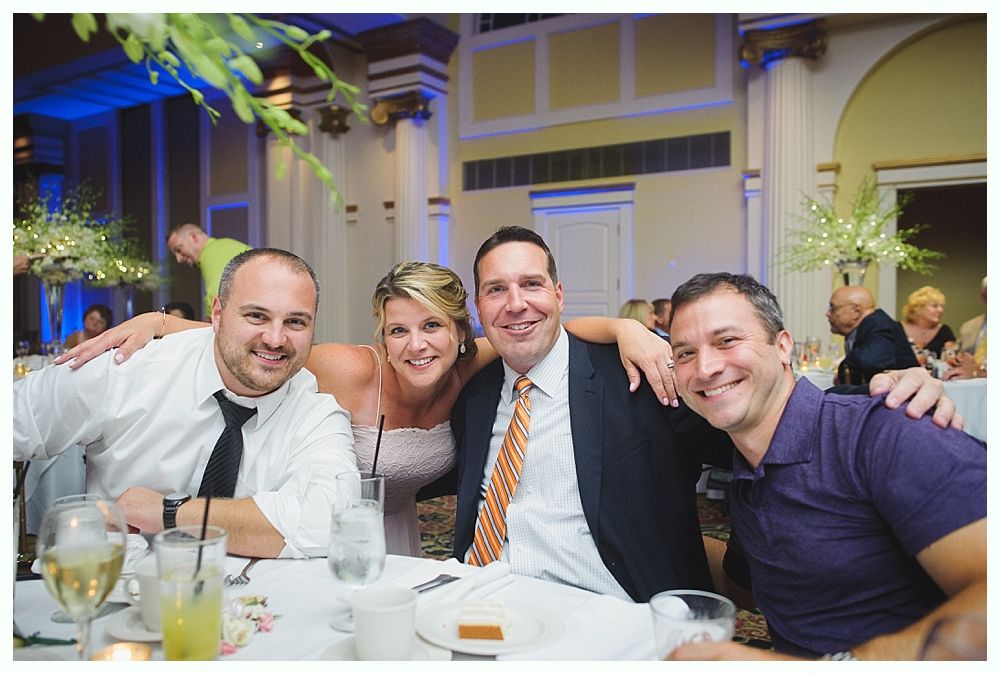 Bride with arms up, making a funny face, groom looking at his hand, cake cutting in background.