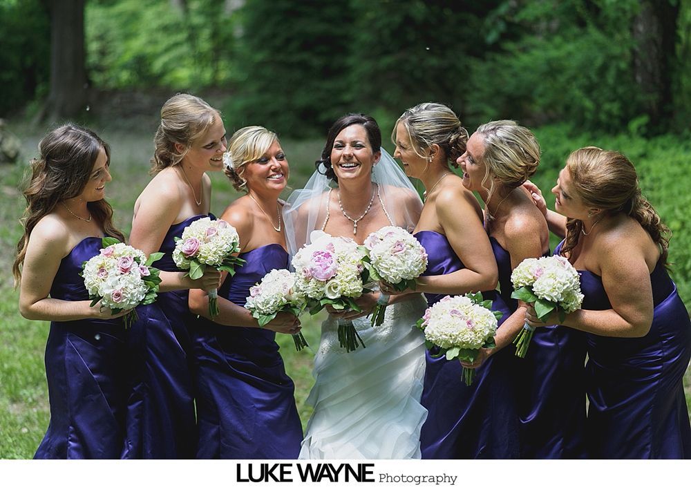 Bridesmaids in pastel dresses hold bouquets, posing outside a building next to a sign for the Hill-Stead Museum.