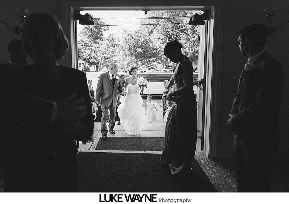 Bride walks down the aisle with her father at a wedding. Guests seated on either side of the aisle. Venue decorated with hanging lanterns.