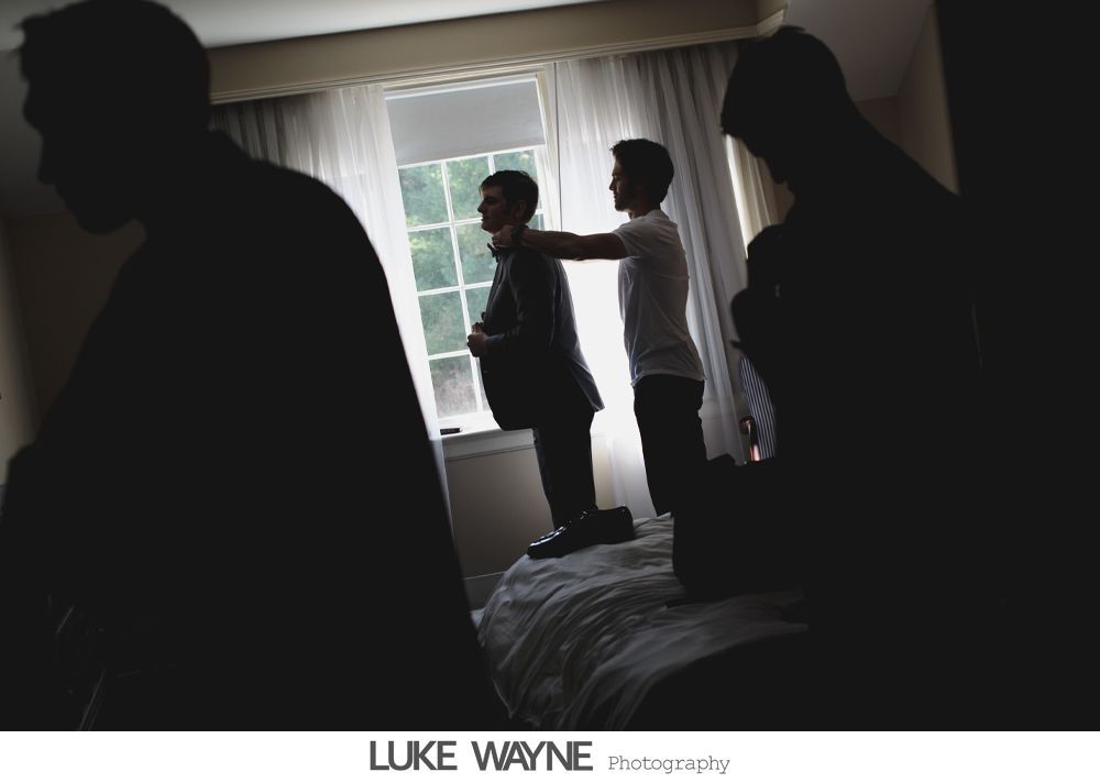 Silhouetted men helping each other with suits in a bright room by a window.