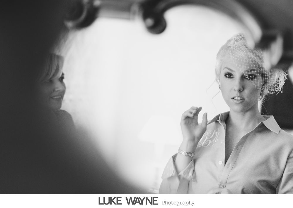 Woman in a button-down shirt adjusts a headpiece, looking at her reflection in a mirror. Black and white photo.