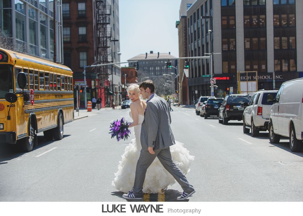 Bride and groom cross a city street, yellow school bus and traffic in the background.