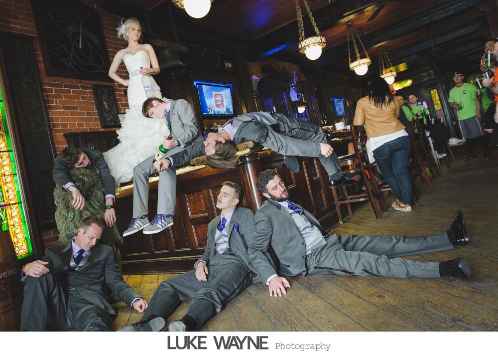 Wedding party posing in bar: bride on bar, groomsmen sprawled on floor, expressions of exhaustion.