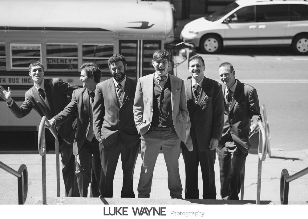 Six men in suits pose on steps by a street. One man has arms open, others smile or look at the camera. A bus and car are in the background.