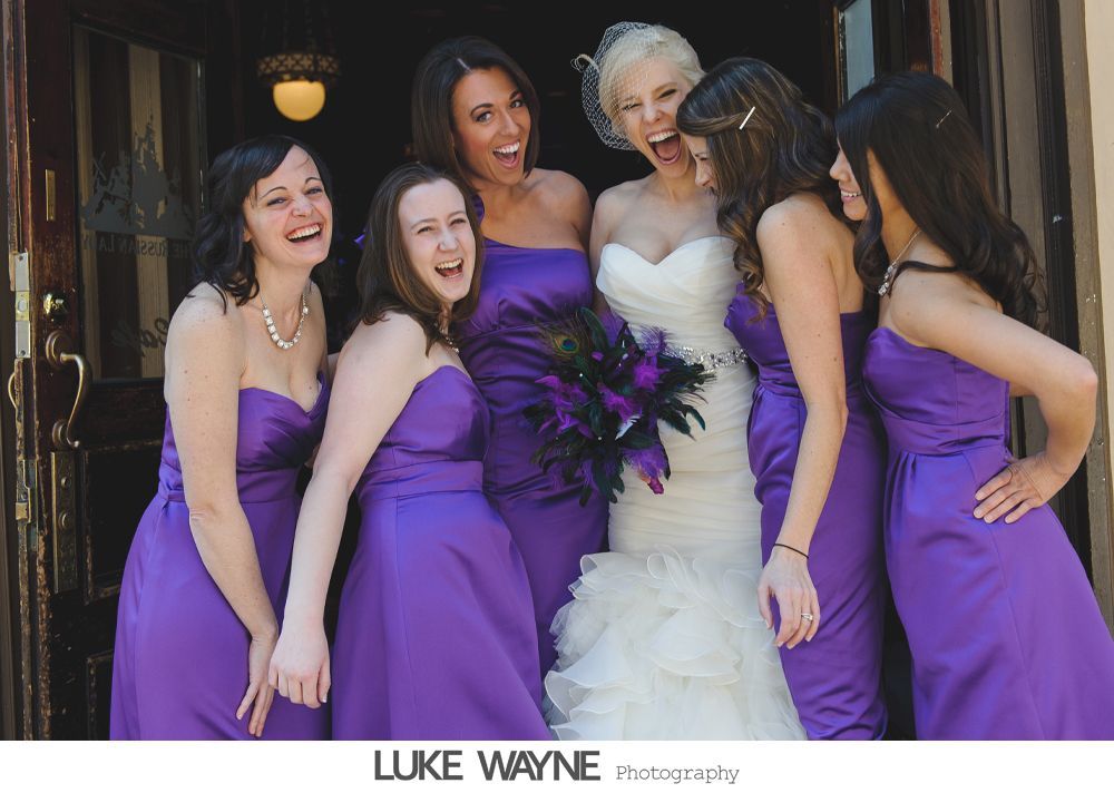 Bride in white dress laughs with bridesmaids in purple gowns.
