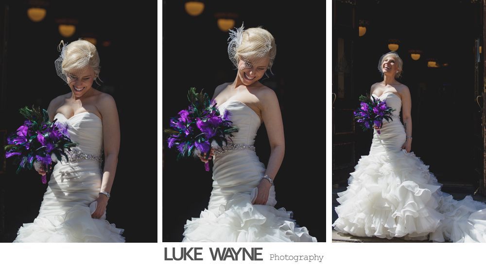 Bride in strapless white dress holding purple flowers, smiling. Dark background with archway.