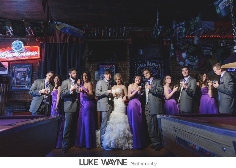 Wedding party toasting with champagne in a dimly lit bar, bride and groom center. Purple dresses, grey suits.