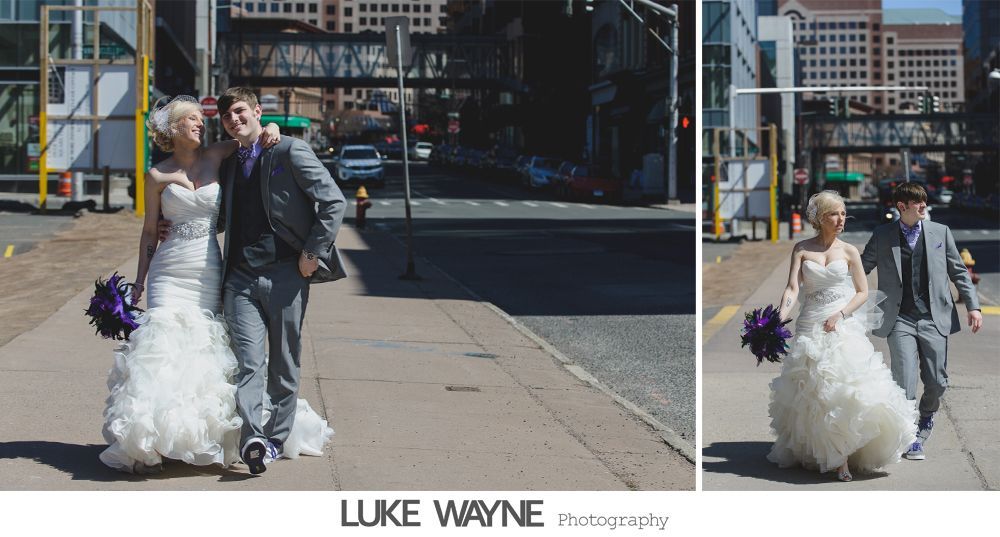 Bride and groom walk down sidewalk, groom smiles, bride holds purple bouquet, city background.