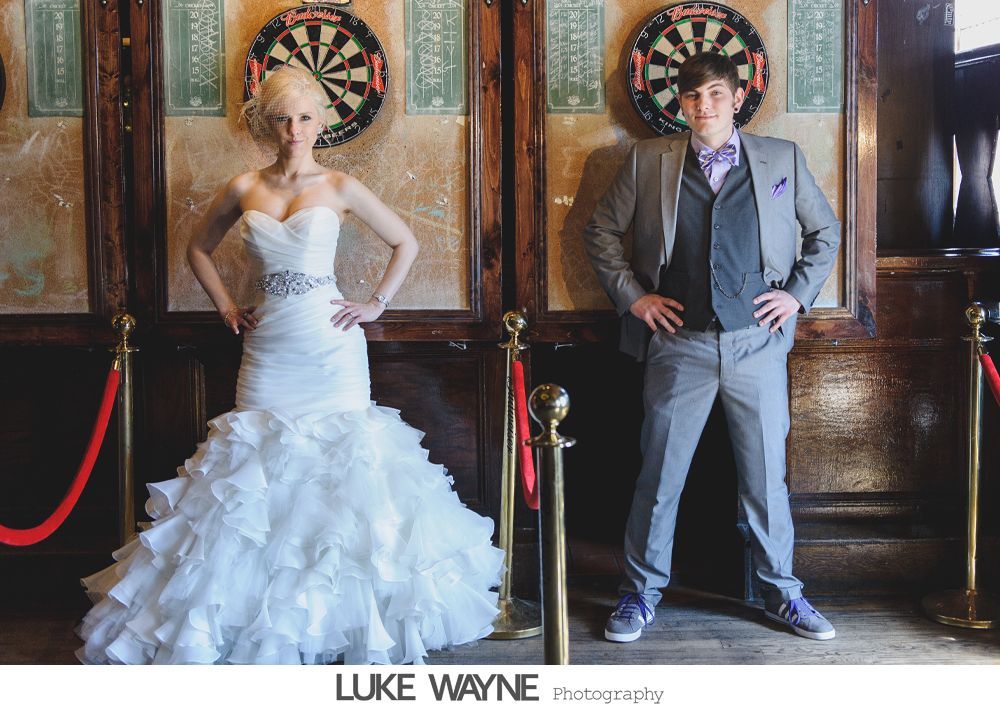 Bride and groom pose near dart boards. Bride in white dress, groom in suit. Indoor setting, red velvet ropes.