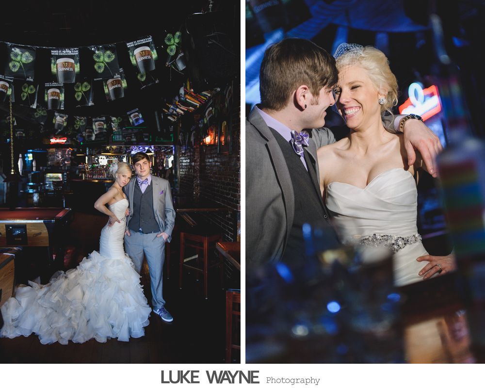 Couple in wedding attire posing in a bar. The woman's dress is white, the man's suit grey, both smiling.