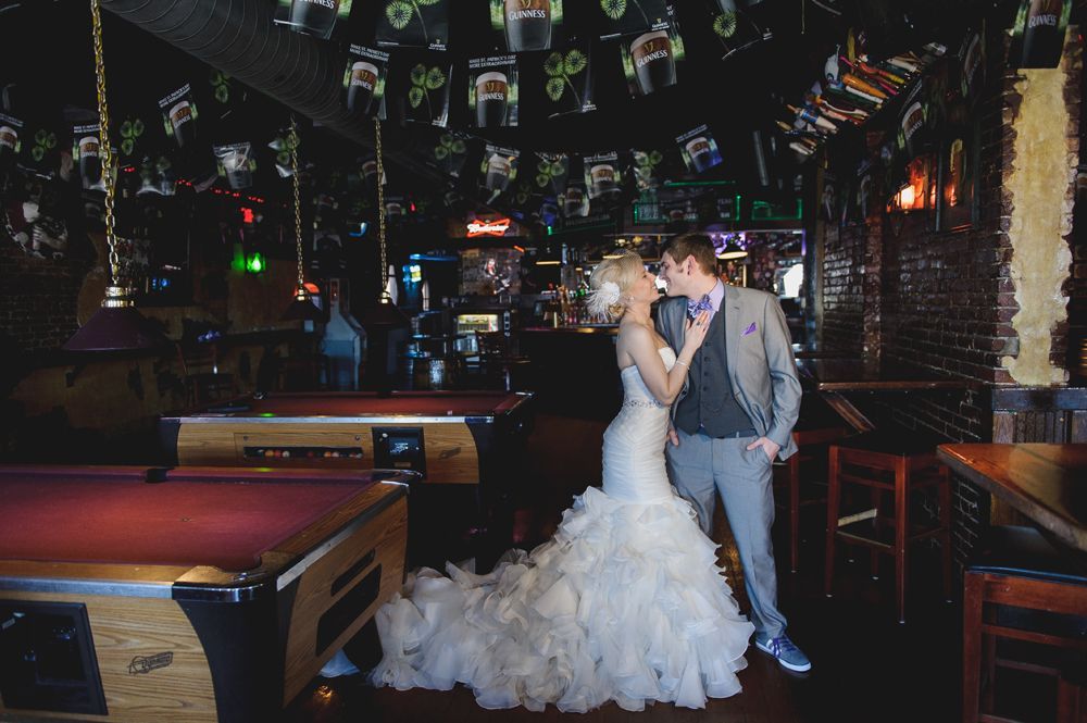 Bride and groom in wedding attire kissing in a dimly lit bar, next to a pool table.