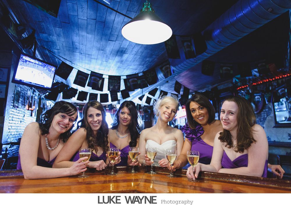 Bride and bridesmaids in purple dresses at a bar, toasting with drinks.