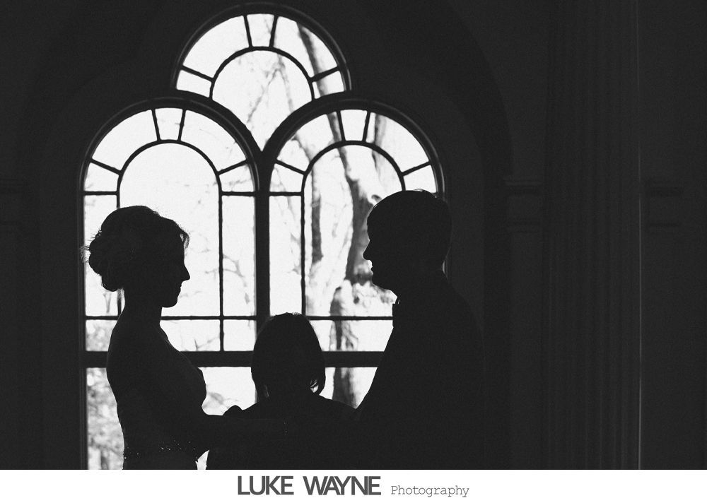 Silhouetted wedding couple and officiant in front of an arched window.