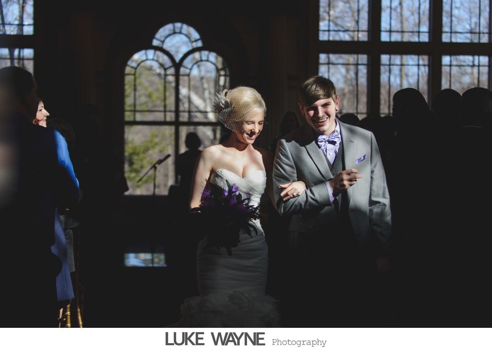 Bride and groom exit a building, holding arms, smiling. Bright sun streams through windows.