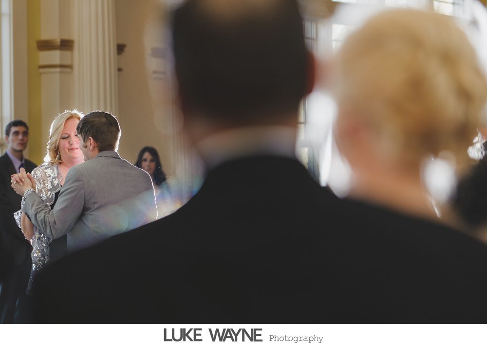 People dancing in a sunlit room; a couple embraces on the dance floor, others watch.