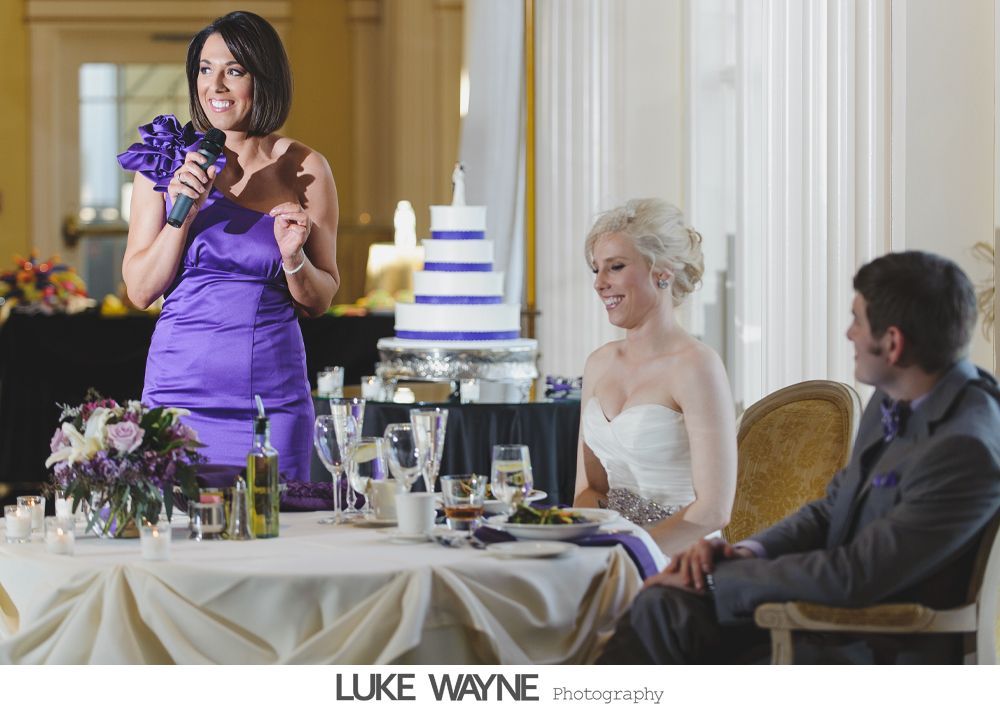 Woman in purple dress giving a speech at a wedding, bride and groom seated at table.