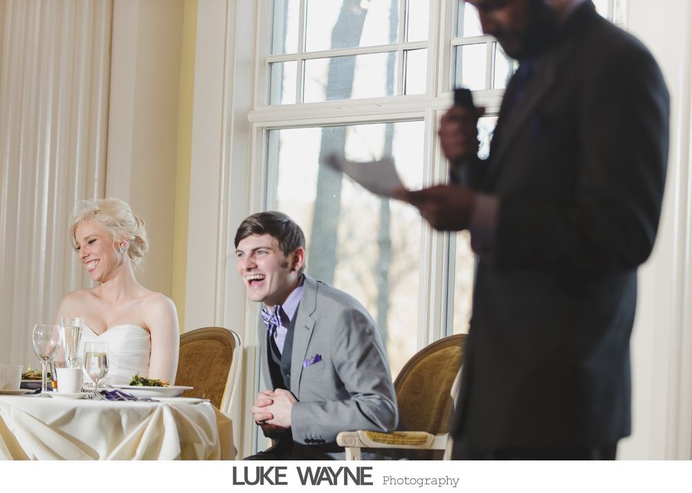 Bride and groom laughing at a wedding reception while a man gives a speech.