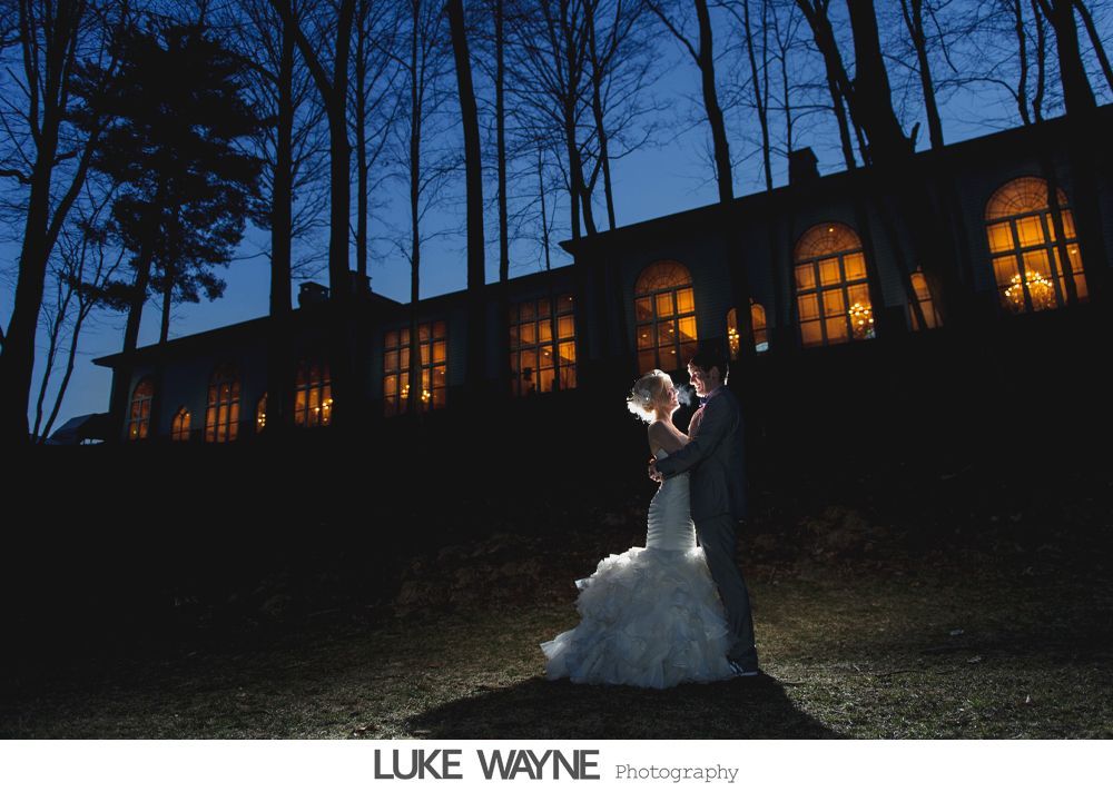 Bride and groom embrace, lit by spotlight, in front of a building with glowing windows at night.