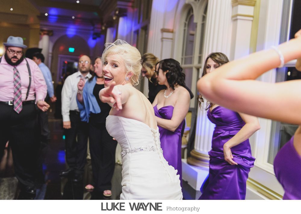 Bride dancing at a wedding reception; wearing a white gown, pointing; bridesmaids in purple dresses.