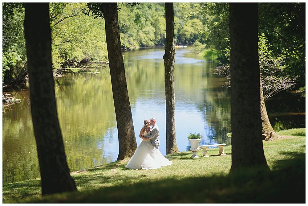 Couple embraces in wedding attire by a river framed by trees; sunlight, greenery, and bench present.