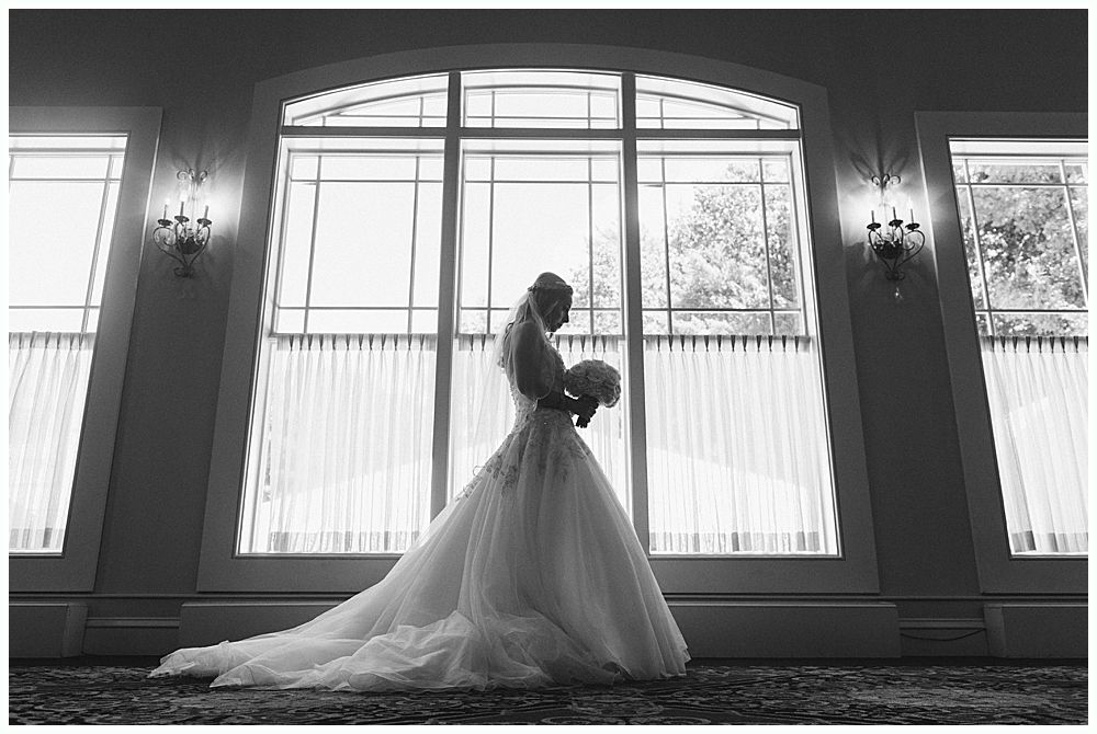 Bride in silhouette holding bouquet, standing before large window.