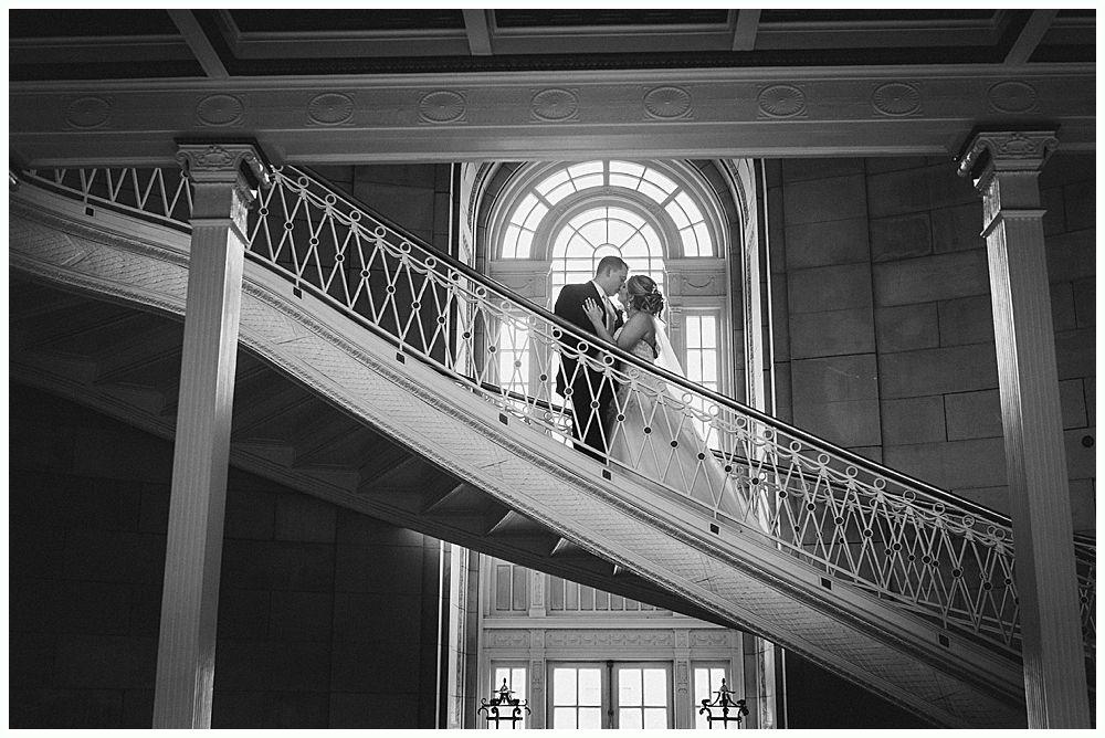 Bride and groom kissing on a grand staircase with arched window backdrop. Black and white.