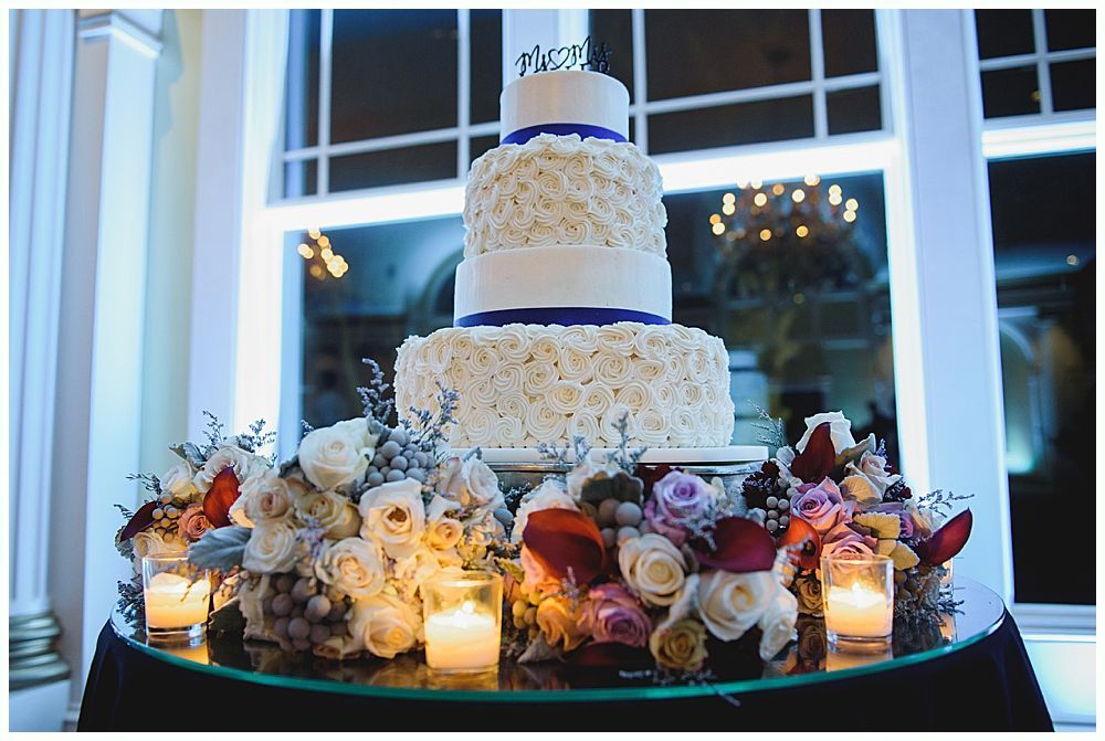 Four-tiered wedding cake with white frosting and blue ribbon, surrounded by flowers and candles.
