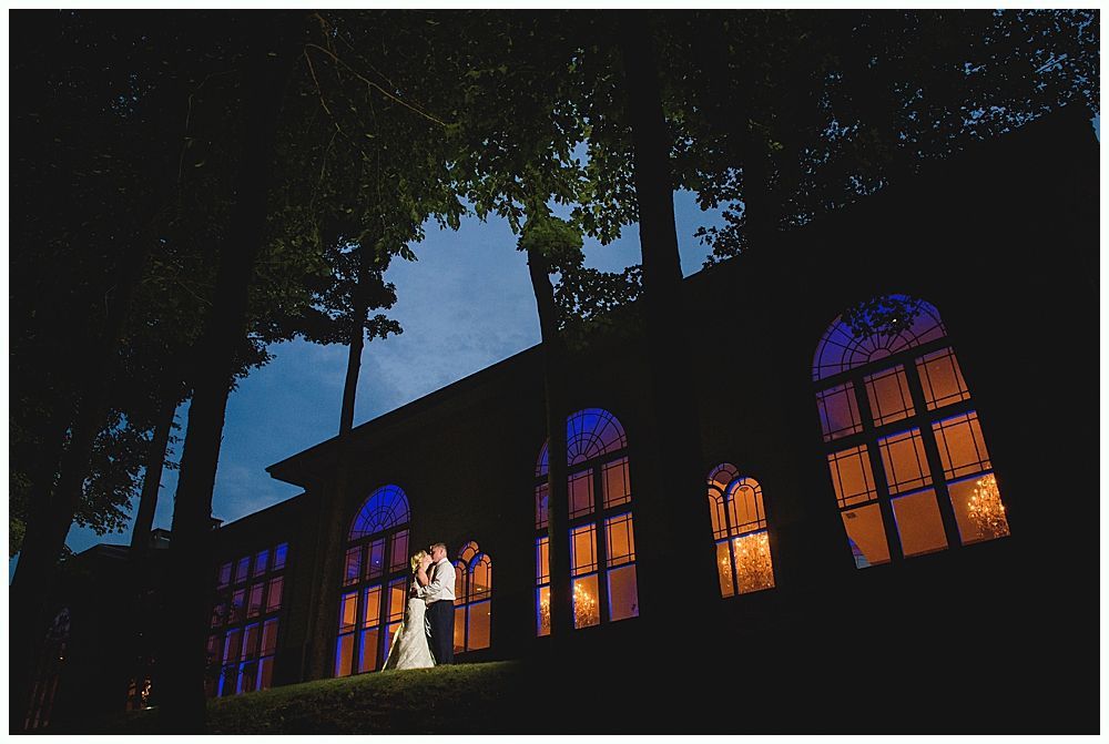 Couple in wedding attire stands before arched windows, illuminated by blue and orange light, at dusk.