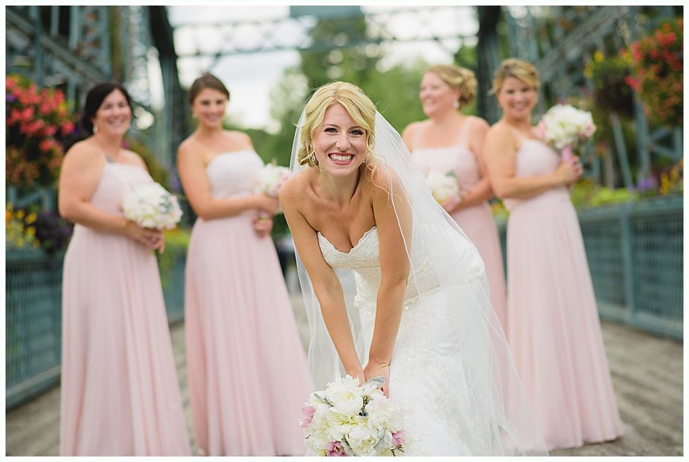 Bride in white gown smiles, surrounded by bridesmaids in pink dresses on a bridge.