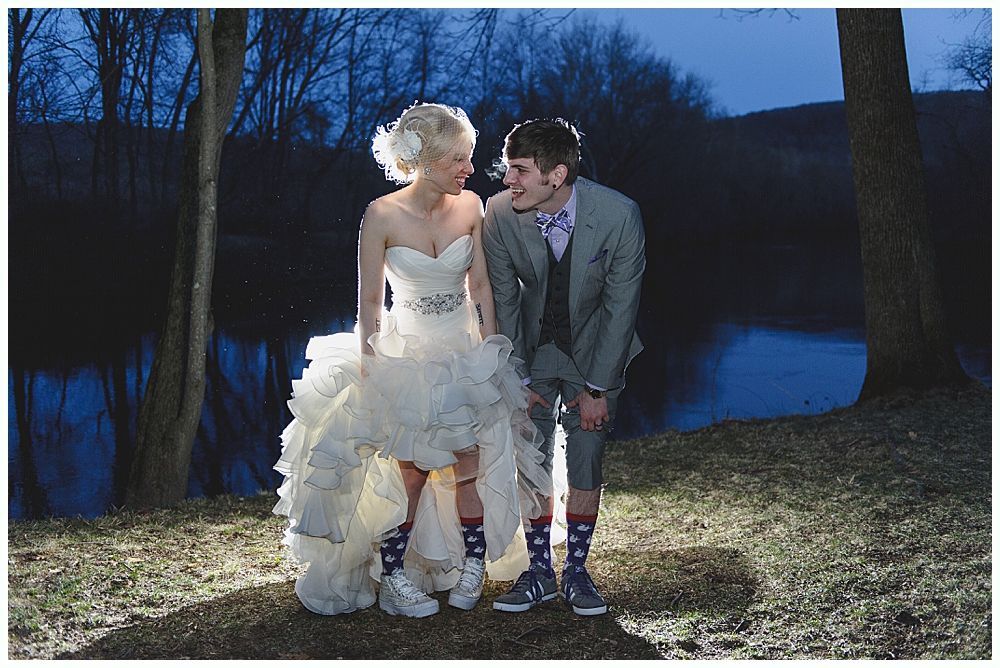 Bride and groom in wedding attire, shoes visible, standing near water. Nighttime, trees.