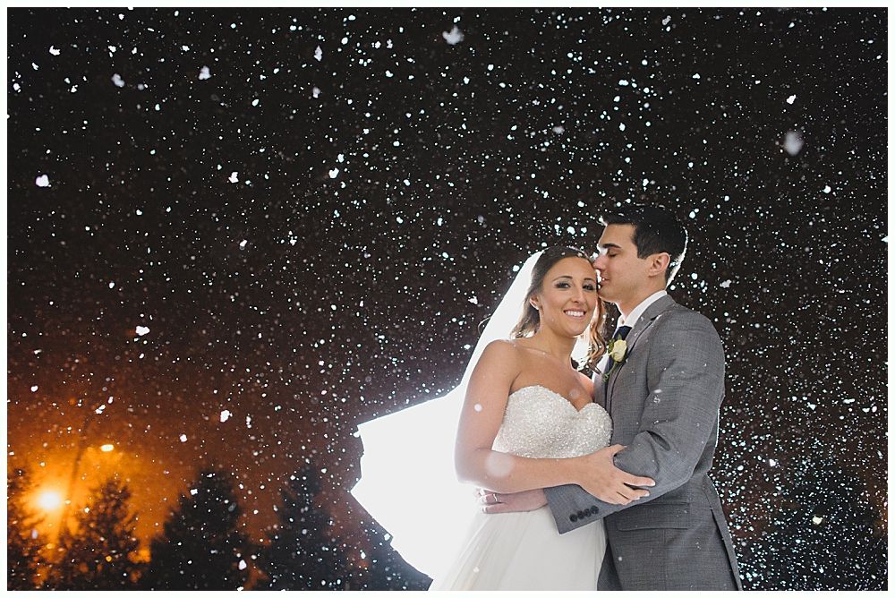Newlyweds embrace under falling snow at night. Bride in white dress, groom in grey suit, smiling.