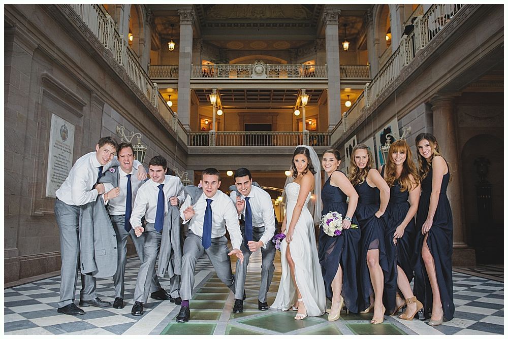 Wedding party poses in a grand hall: bride with bridesmaids, groomsmen leaning forward.