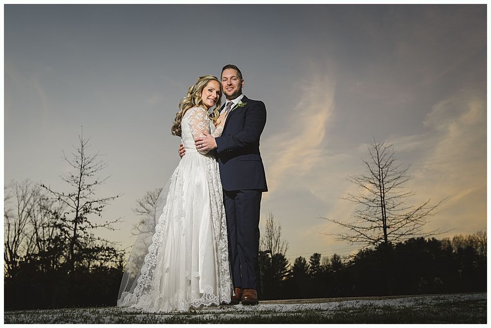 Wedding couple embraces, posing against a dusky sky with bare trees. The bride wears a lace dress, the groom a navy suit.