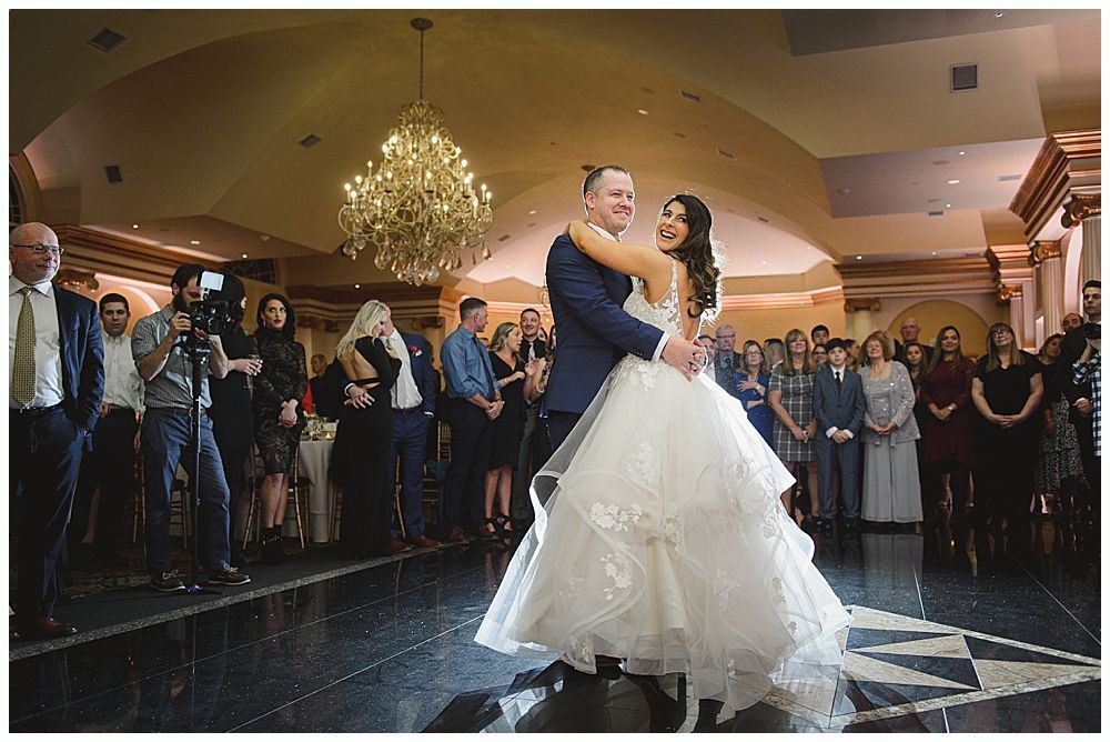 Couple dancing at a wedding reception; chandelier hangs above. Guests watch in background.