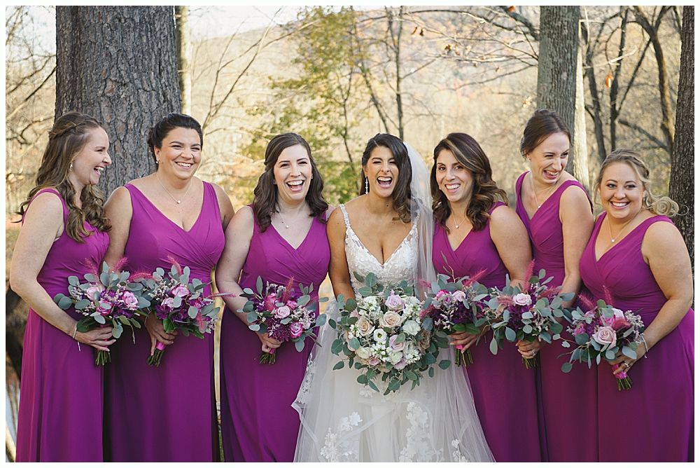 Bride and bridesmaids in magenta dresses laugh outdoors, holding bouquets.