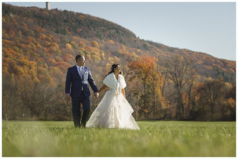 Bride and groom holding hands, walking in a grassy field with fall foliage and a mountain in the background.