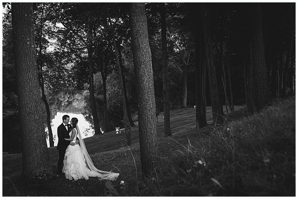 Bride and groom embrace in a forest, she in a gown with a long veil, he in a suit. Black and white.