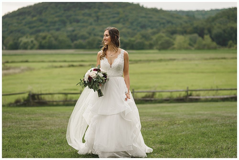 Bride in white dress holding bouquet, standing in field with wooded hill in background.