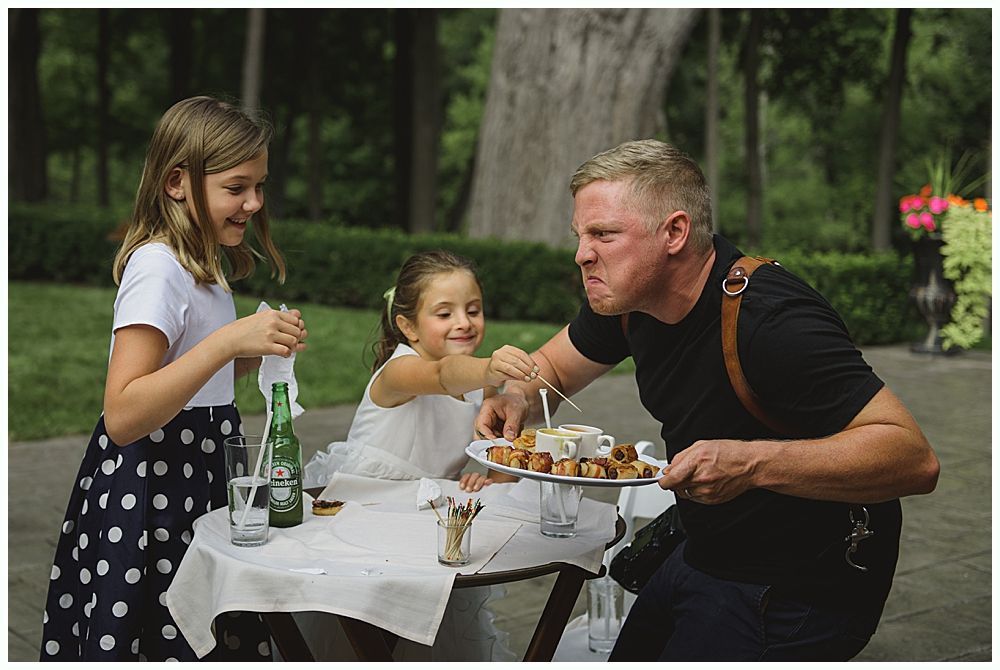 Man eats food from a plate while two girls watch; outdoors at a table.
