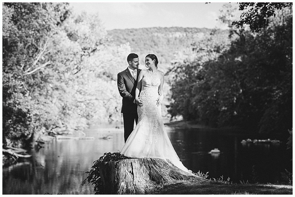 Wedding couple stands holding hands, smiling, on a tree stump by a river, surrounded by trees.